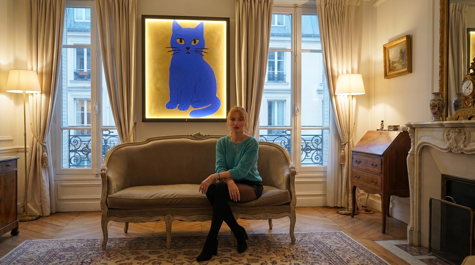Woman sitting on a beige sofa in a stylish living room with a blue cat artwork on the wall.
