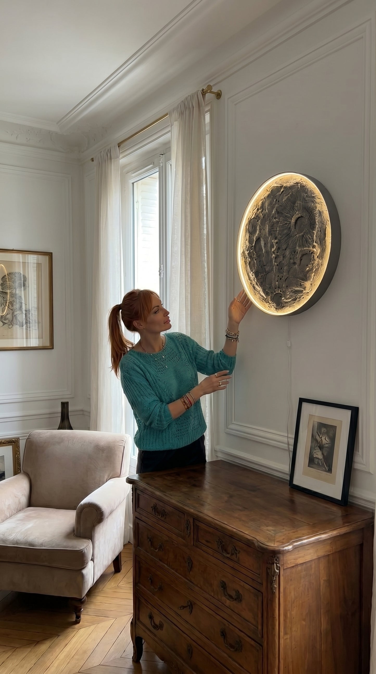 Woman adjusting a large round mirror on a wall in a room with a wooden dresser and framed pictures.
